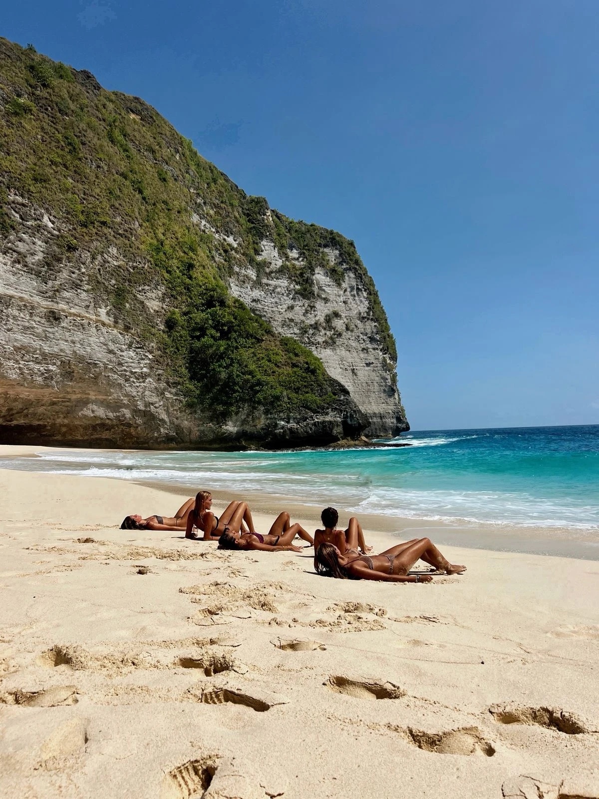 Friends sunbathing on a white-sand beach below a green cliff in Bali